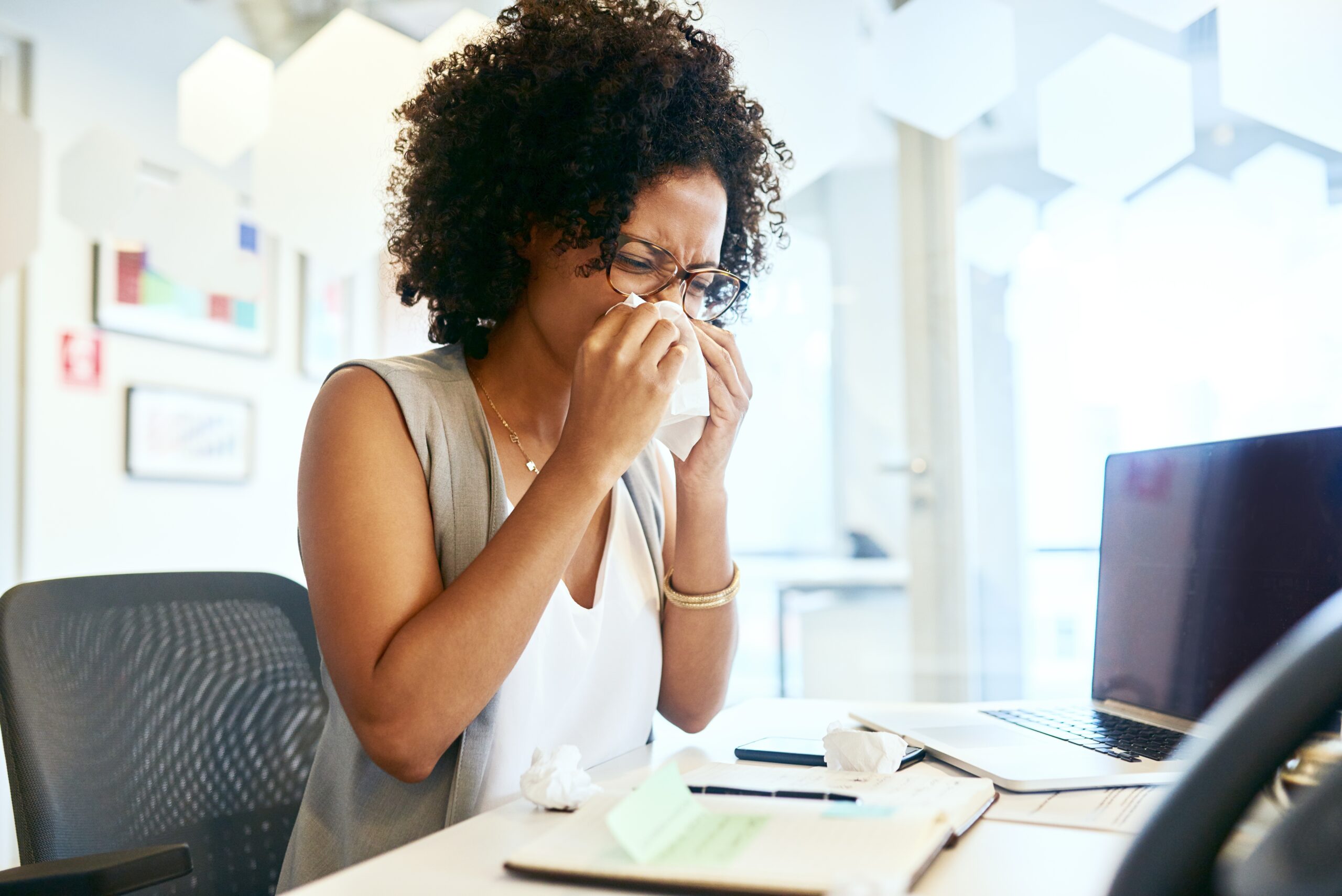 Young woman sat at her desk blowing her nose.