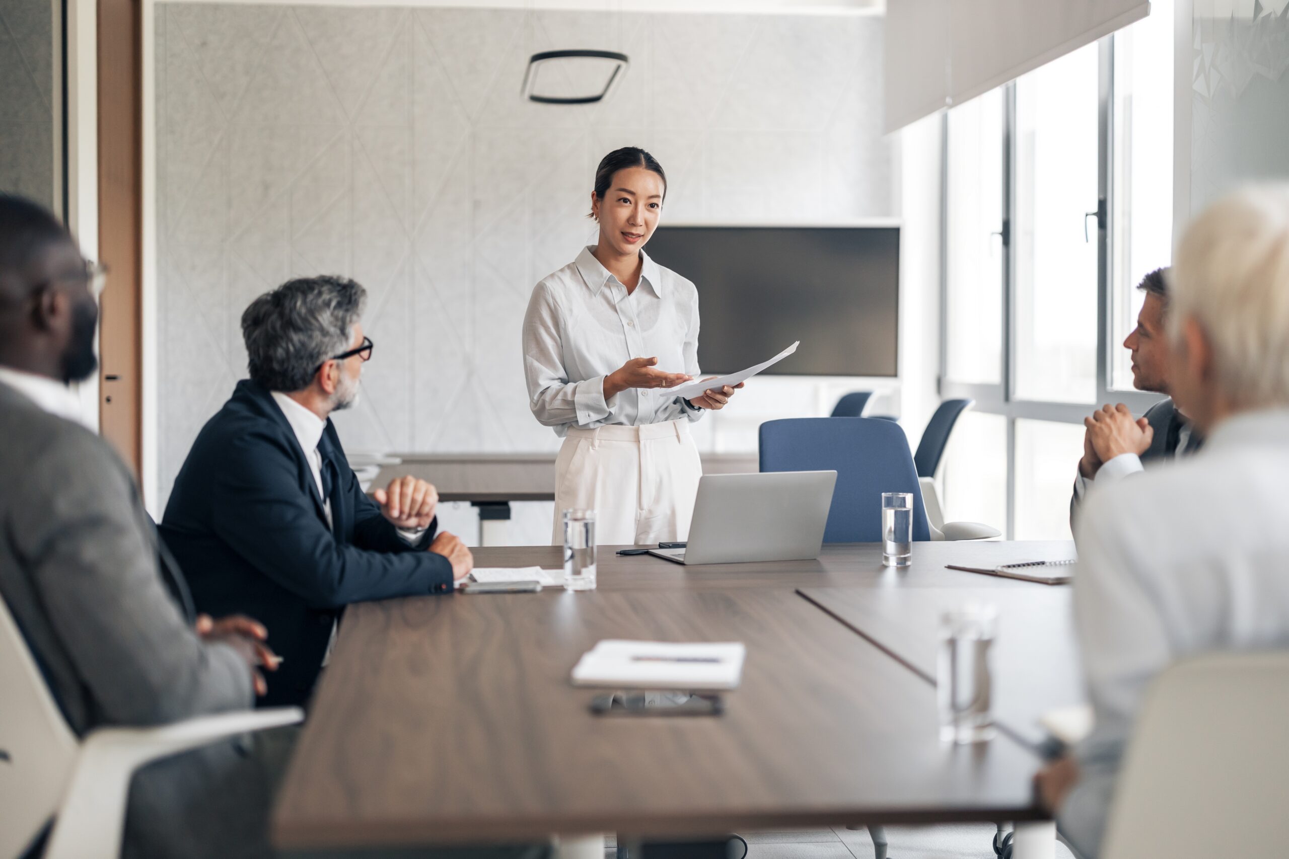Businesswoman presenting reports to diverse colleagues in a modern office boardroom standing by a laptop
